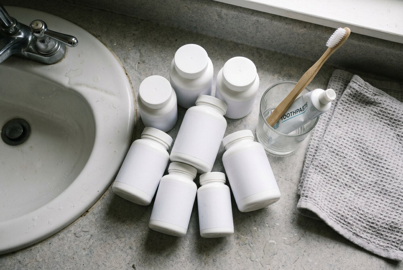 A cluttered pile of separate supplement bottles on a bathroom bench by the sink